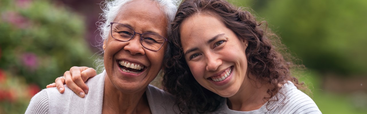 Two women, one a younger adult and the other an older adult, stand arm in arm smiling in a park