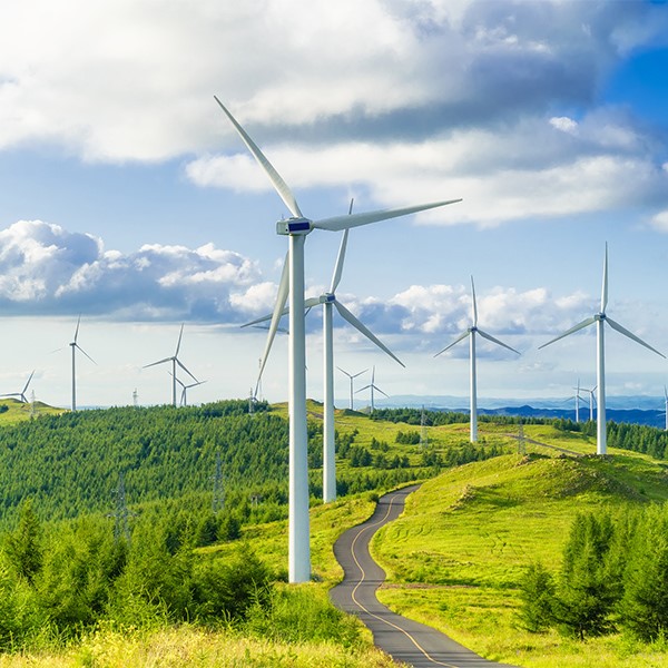 Wind energy farm with a blue sky in the background and a winding road going through rolling green hills.ll