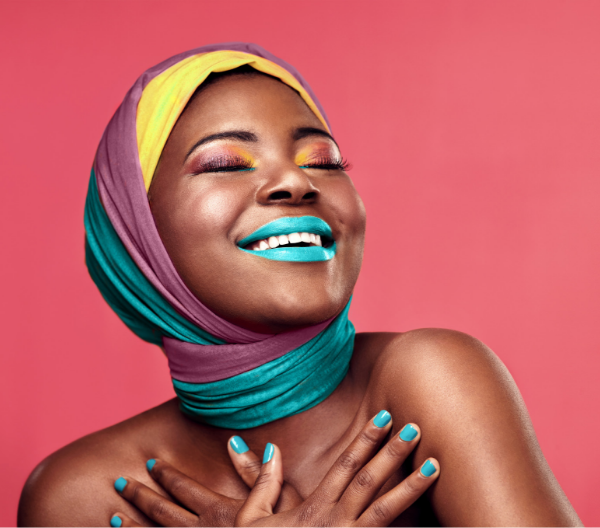 Woman wearing a colorful headscarf, wearing bright blue lipstick, laughing in front of a coral background